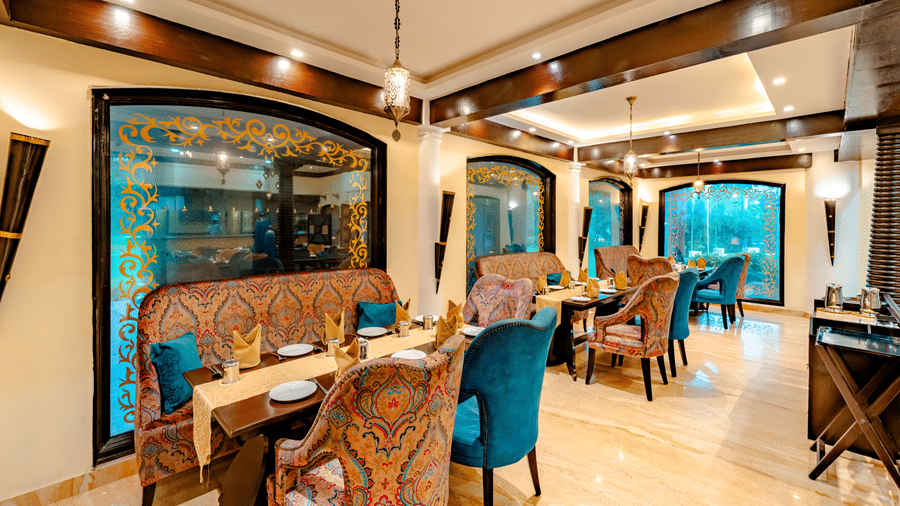 An interior view of Za-aiqa, Noor-Us-Sabah Palace, Bhopal, showing an empty dining hall with set tables, decorative arched windows, and a mix of patterned and solid teal chairs.