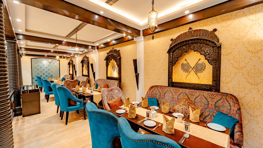 The sophisticated main dining area of Za-aiqa, featuring golden damask wallpaper, decorative wooden wall accents with musket motifs, and plush teal velvet chairs.