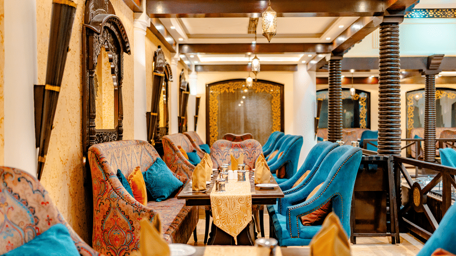 A close-up shot of the seating at Za-aiqa, found at Noor-Us-Sabah Palace, Bhopal, showcasing plush teal and patterned armchairs arranged around long wooden tables with traditional architectural accents in the background.