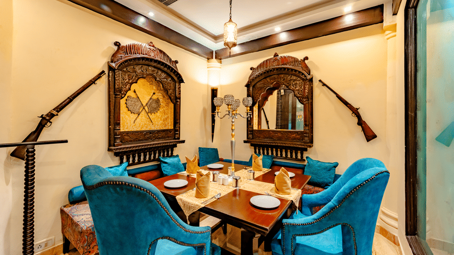 A cosy dining nook at Za-aiqa, located within Noor-Us-Sabah Palace, Bhopal, featuring plush teal armchairs and walls adorned with traditional decor, including antique muskets and carved wooden mirrors.