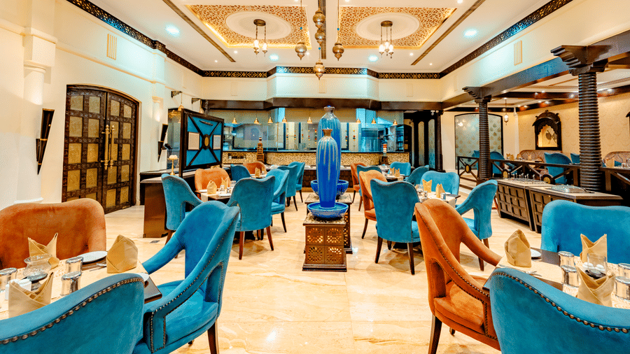 The elegant main dining hall of Za-aiqa at Noor-Us-Sabah Palace, Bhopal, showcasing a mix of vibrant teal and orange velvet seating, polished marble floors, and warm, ornate lighting.