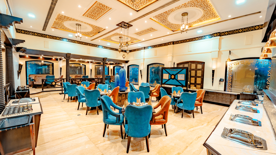 A wide-angle view of the dining hall at Za-aiqa, Noor-Us-Sabah Palace, Bhopal, featuring vibrant teal and orange velvet chairs and a polished marble floor.