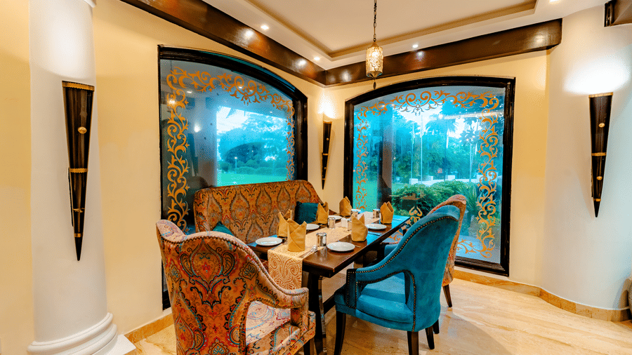 An intimate dining corner at Za-aiqa, Noor-Us-Sabah Palace, Bhopal, featuring plush teal and patterned chairs beside large decorative windows.
