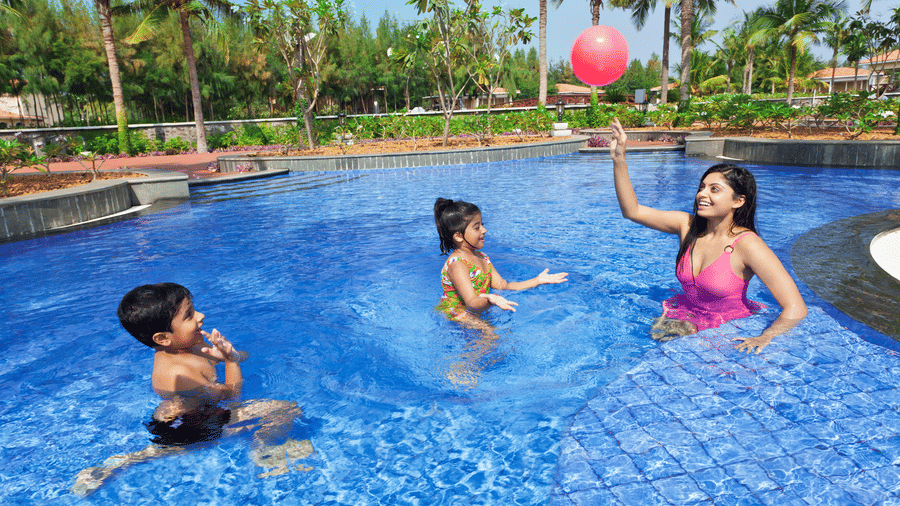 A mother and her 2 children playing in a swimming pool at OCEAN SPRAY resort, with palm trees and garden in the background.