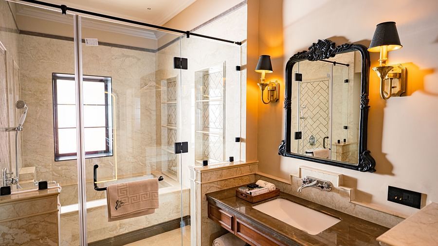 Spacious and elegant hotel bathroom featuring a large glass-enclosed shower area with a built-in bench, beige marble walls, a vanity with a marble countertop and black-framed mirror, golden wall sconces, rolled towels, and a tray with complimentary toiletr