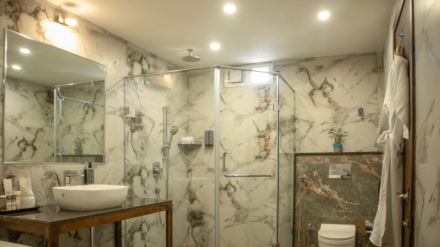A modern Bathroom of a Suite at Papaya Tree Hotels with a wall-mounted mirror, a ceramic sink, a separate shower cubicle with ample amount of lighting.