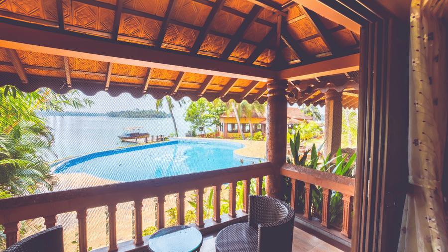 Balcony with tiled roof, two black chairs, and a small table, overlooking water and trees at Paradise Lagoon Resort, Udupi.
