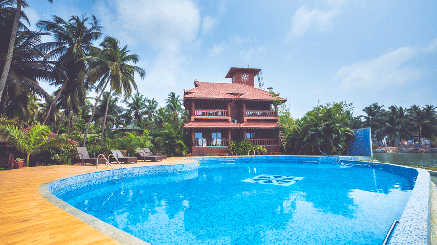 Swimming pool surrounded by palm trees, with a red-roofed house in the background at Paradise Lagoon Resort, Udupi.
