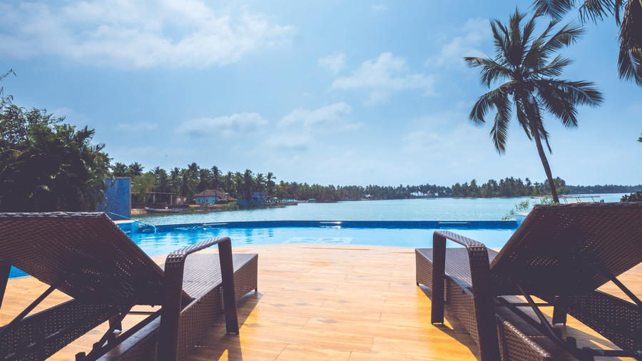 Poolside loungers facing an infinity pool with palm trees, water view, and blue sky in the background at Paradise Lagoon Resort, Udupi.