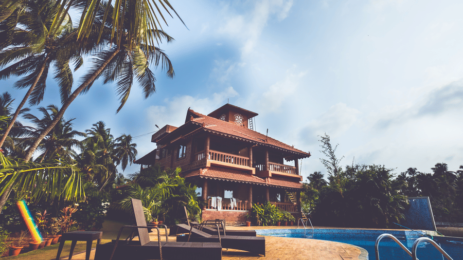 Red-roofed building with steps leading to a pool, surrounded by loungers & palm trees under blue sky at Paradise Lagoon Resort, Udupi.