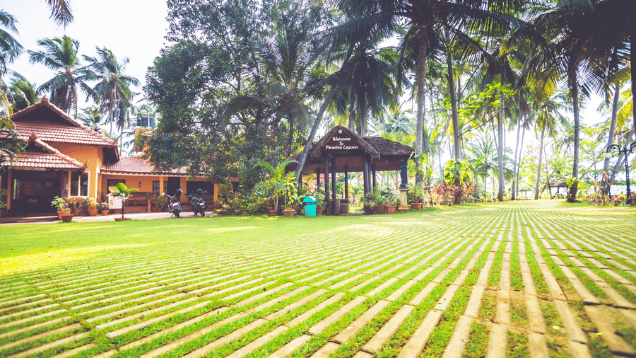 Lawn with palm trees, tiled pathways, and red-roofed building surrounded by greenery at Paradise Lagoon Resort, Udupi.