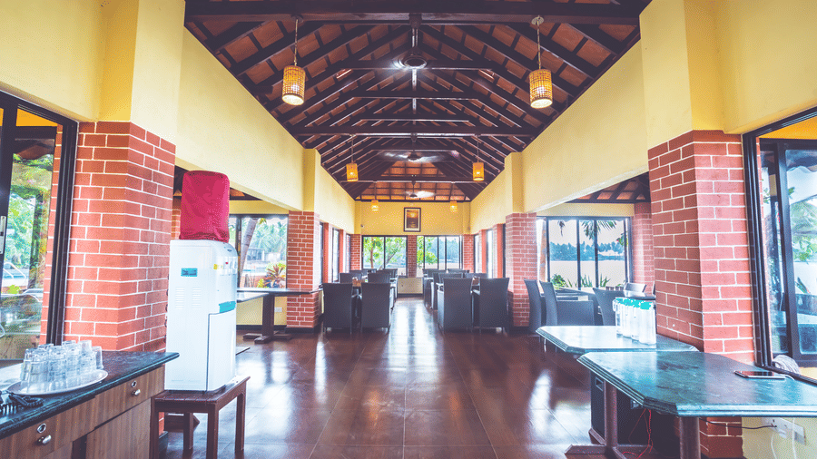 Dining area with wooden tables and chairs, brick pillars, tiled floor, and sloped ceiling with lights at Paradise Lagoon Resort, Udupi.