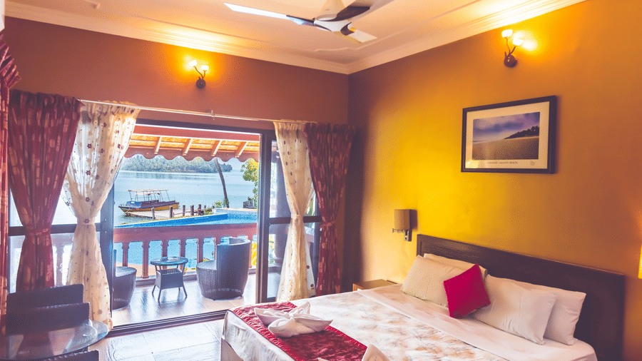 Room with a queen size bed, ceiling fan, framed picture, and view of the river through the window at Paradise Lagoon Resort, Udupi.