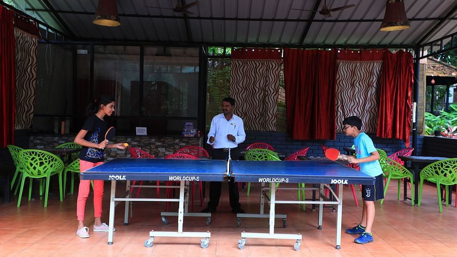 Two people playing table tennis and a man watching them at Parampara Resort & Spa, Kushalnagar.