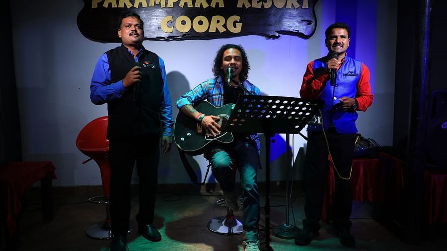 A man holding a guitar and two men holding microphones at Parampara Resort & Spa, Kushalnagar.