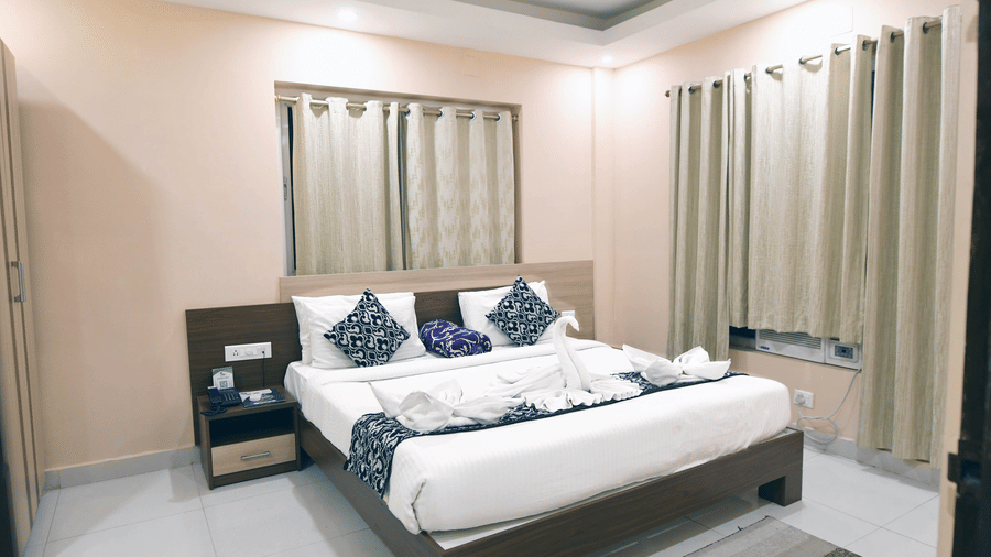 Bedroom of an Executive Suite at Pipul Hotel And Resort, Puri, featuring a plush bed with decorative towel art, patterned cushions, a nightstand, and draped windows.