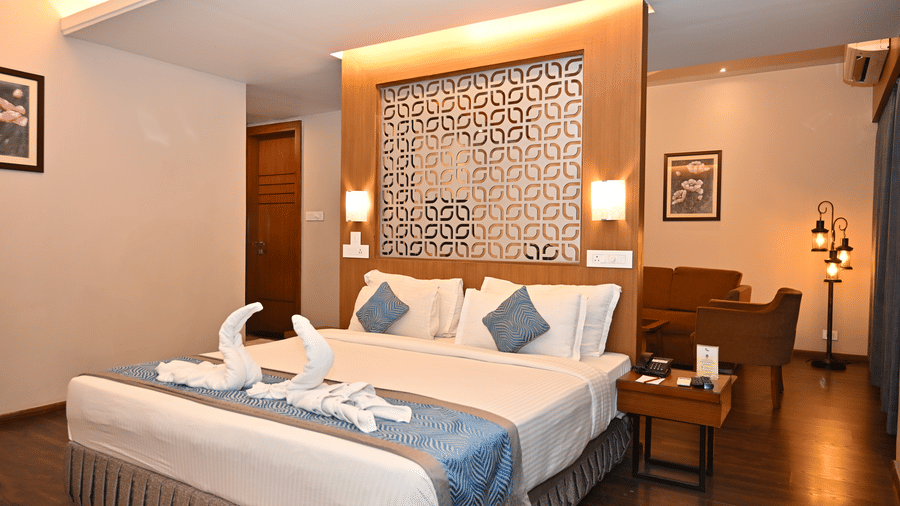 Suite bedroom with a bed, swan-shaped towel art, a patterned wall panel headboard, a bedside lamp and a small seating area near the window.