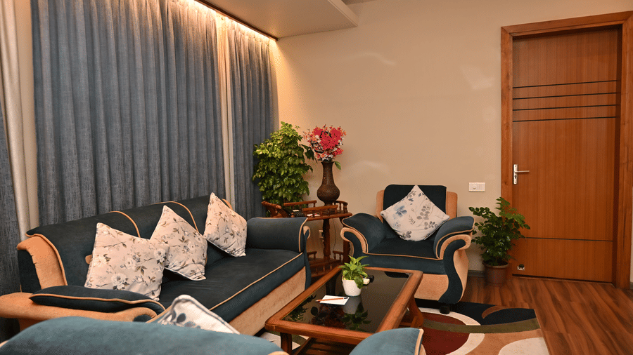Suite living area with a sofa dressed in blue and white cushions, a wooden coffee table, flowers on display and long curtains by the window.