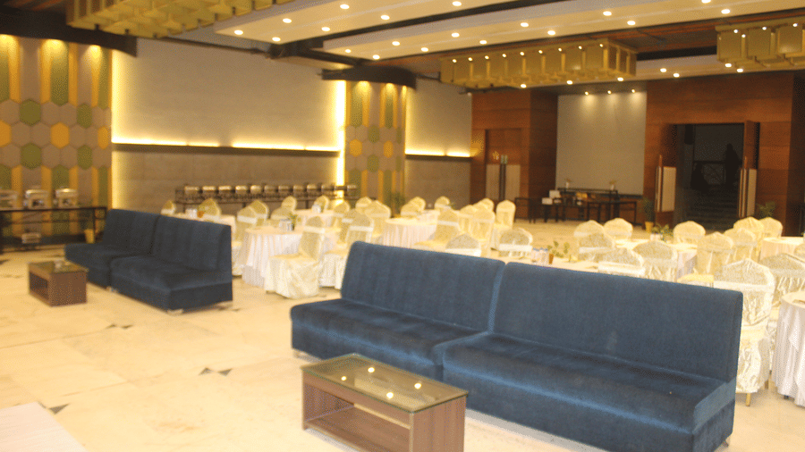 A banquet hall with 2 blue attached sofas, a table, and rows of covered chairs behind with ceiling lights at Pipul Padmaja Premium Hotel & Convention.