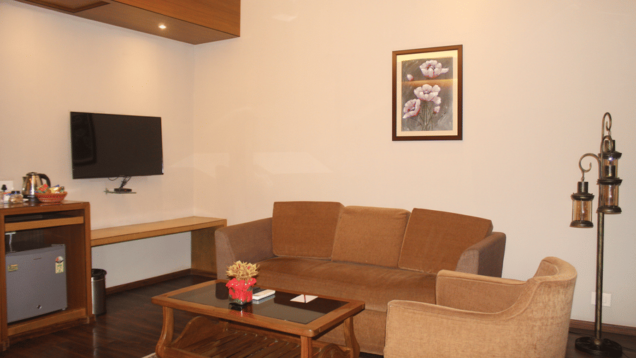 Close-up of a living room with a tan-coloured two-seater sofa, matching armchair, wooden coffee table, wall-mounted television, minifridge, and electric kettle station at Pipul Padmaja Premium Hotel & Convention.