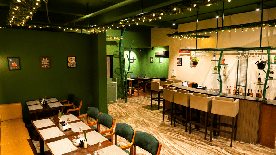  The interior of a casual dining room at Pravasa Hotel, Kolhapur, featuring green walls, simple laid tables, and a breakfast bar area.
