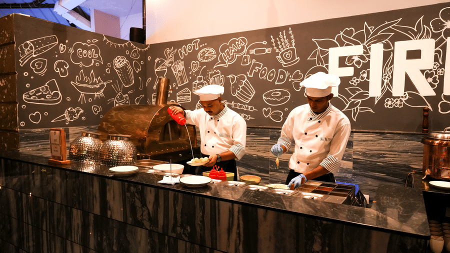  Two chefs preparing food at a live cooking station with a pizza oven and a chalkboard wall in Pravasa Hotel, Kolhapur.