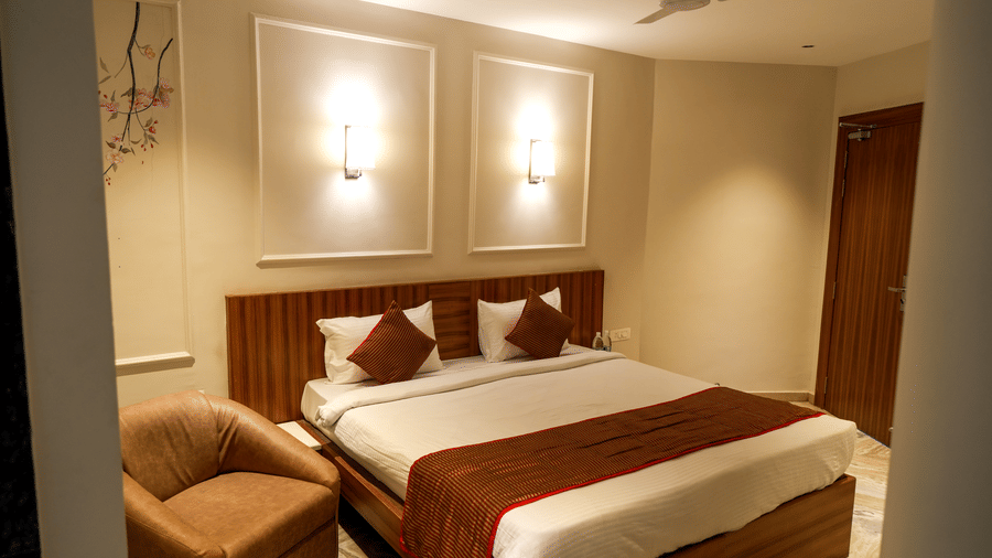  A modern guest bedroom at Pravasa Hotel, Kolhapur, featuring a double bed, subtle wall panel moulding, and a tan armchair. 