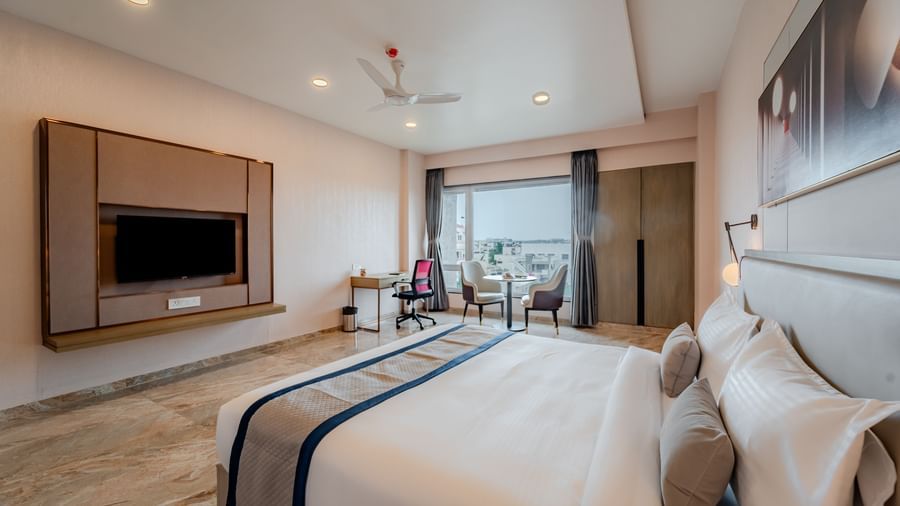  A wide angle shot of a hotel bedroom with a large bed, TV, and a desk, well-lit | Premium Room | Pravasa Hotel, Vijayapura
