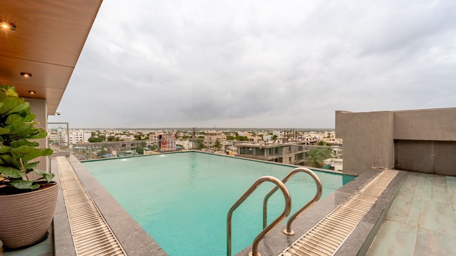 Rooftop swimming pool with clear blue water, surrounded by a tiled deck and some modern outdoor seating, offering a panoramic view of a city under a cloudy sky | Pravasa Hotel Vijayapura