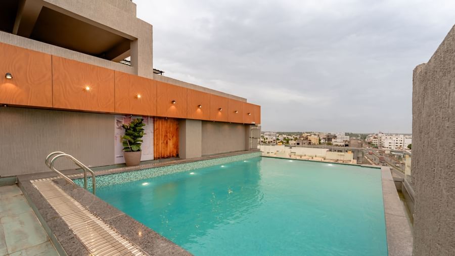 A rooftop swimming pool with clear blue water, surrounded by a tiled deck and some modern outdoor seating, offering a panoramic view of a city under a cloudy sky | Pravasa Hotel Vijayapura