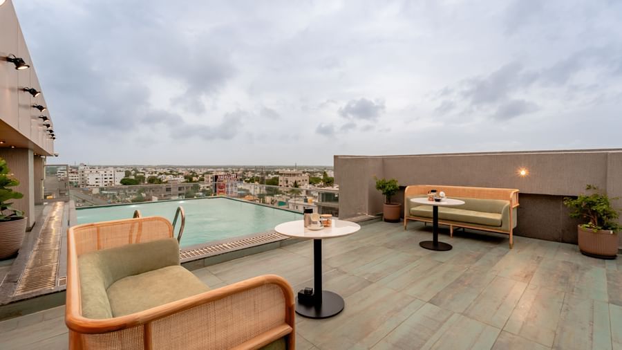 The rooftop swimming pool with clear blue water, surrounded by a tiled deck and some modern outdoor seating, offering a panoramic view of a city under a cloudy sky | Pravasa Hotel Vijayapura