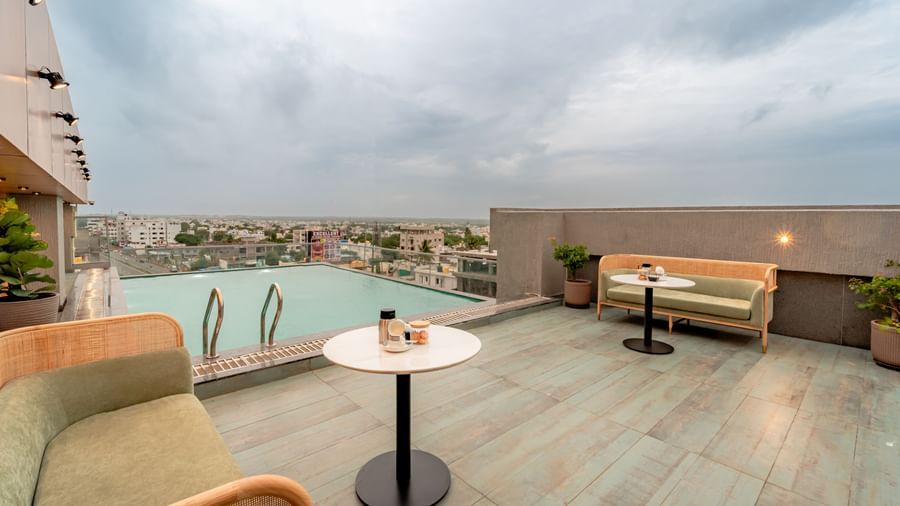 The rooftop swimming pool with clear water, surrounded by a tiled deck and some modern outdoor seating, offering a panoramic view of a city under a cloudy sky | Pravasa Hotel Vijayapura