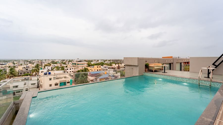 A wide shot of a rooftop swimming pool with clear blue water, surrounded by a tiled deck and some modern outdoor seating, offering a panoramic view of a city under a cloudy sky | Pravasa Hotel Vijayapura