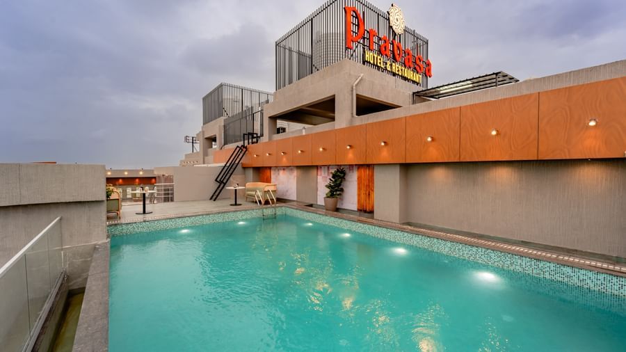 A rooftop swimming pool surrounded by a tiled deck and some modern outdoor seating, offering a panoramic view of a city under a cloudy sky | Pravasa Hotel Vijaypura