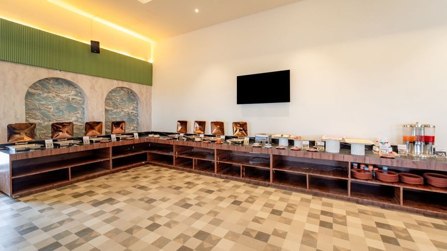 Long buffet counter with an assortment of breakfast items and a TV screen | Pravasa Hotel, Vijayapura