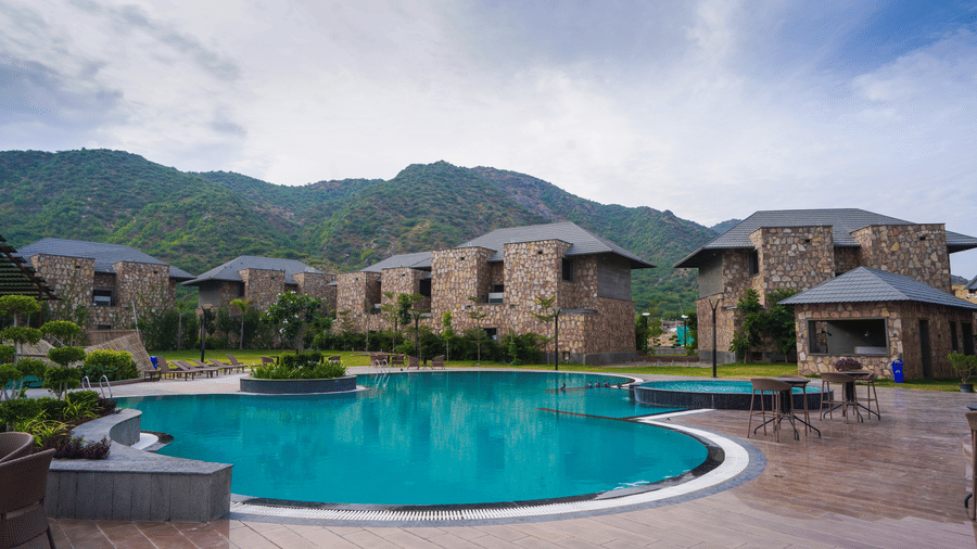 A swimming pool reflects the resort buildings. The surrounding area has view of several holiday villas with a view of the mountains.
