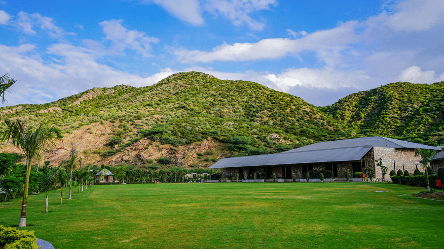 A large, modern resort building sits on a green lawn with a backdrop of lush, green hills under a bright blue, cloudy sky.