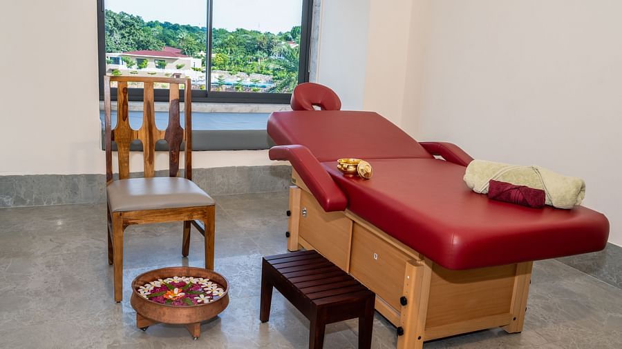 An overview of a massage room featuring a bed, a chair, a small stool, a bowl, and some spa equipment - Rajbagh Wellness.
