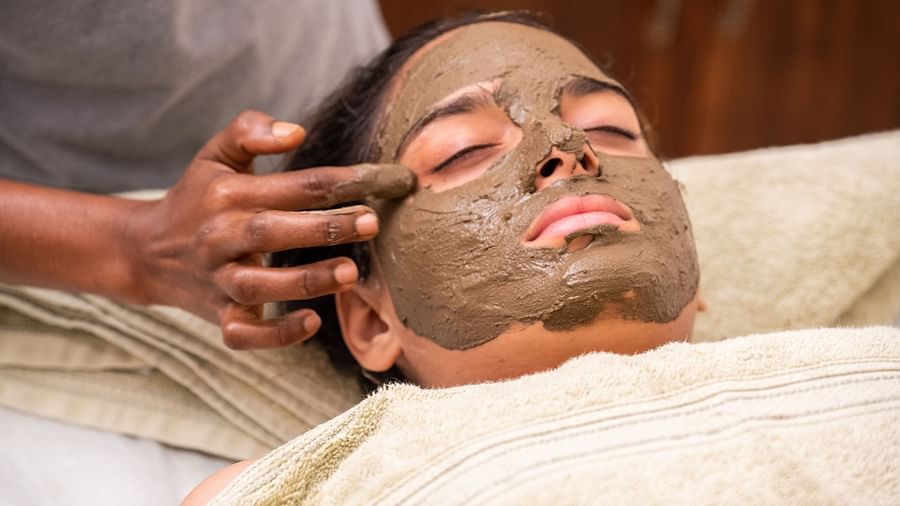 An image of a person applying some paste to a woman's face who is laying on bed - Rajbagh Wellness