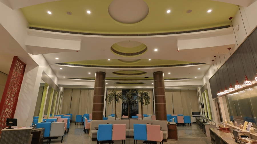 A modern dining hall features a high ceiling, colourful pink and blue chairs, and a long buffet counter at Ramada by Wyndham Bodh Gaya Mahabodhi.