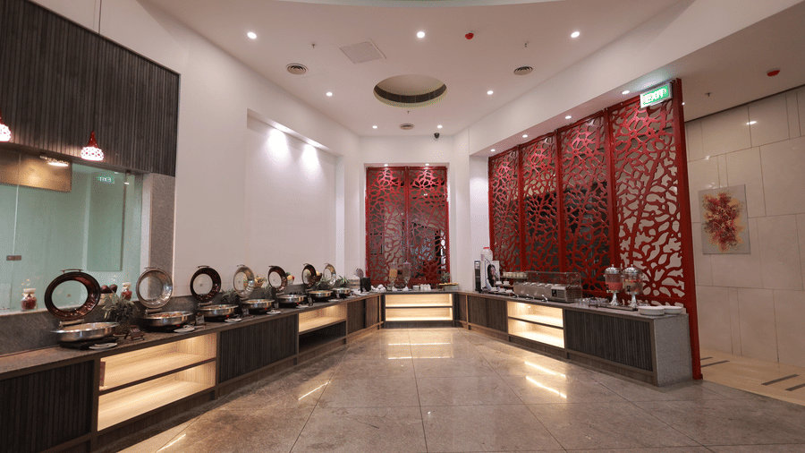 A long, modern buffet area featuring multiple hot and cold serving stations with a decorative red screen wall at Ramada by Wyndham Bodh Gaya Mahabodhi.