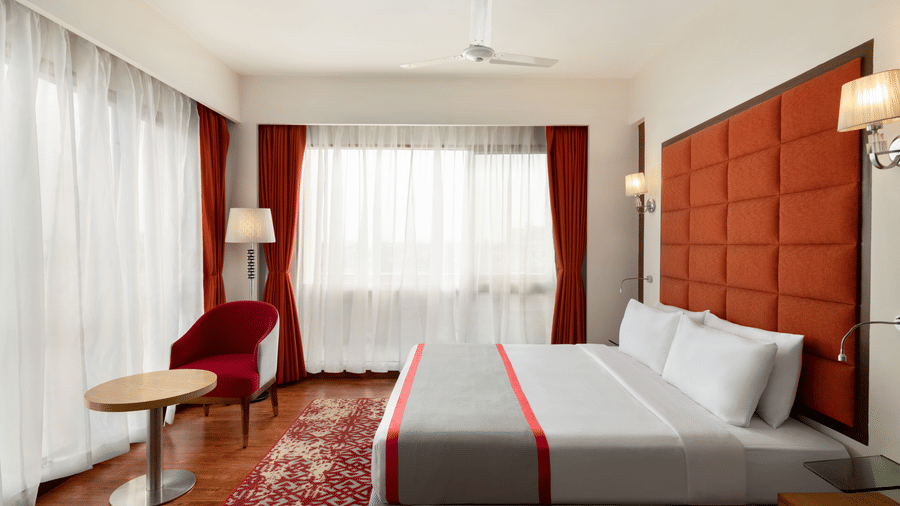 King Suite at Ramada by Wyndham Bodh Gaya Mahabodhi featuring a plush king size bed, a decorative headboard, a small table and a chair, and elegant modern lighting.