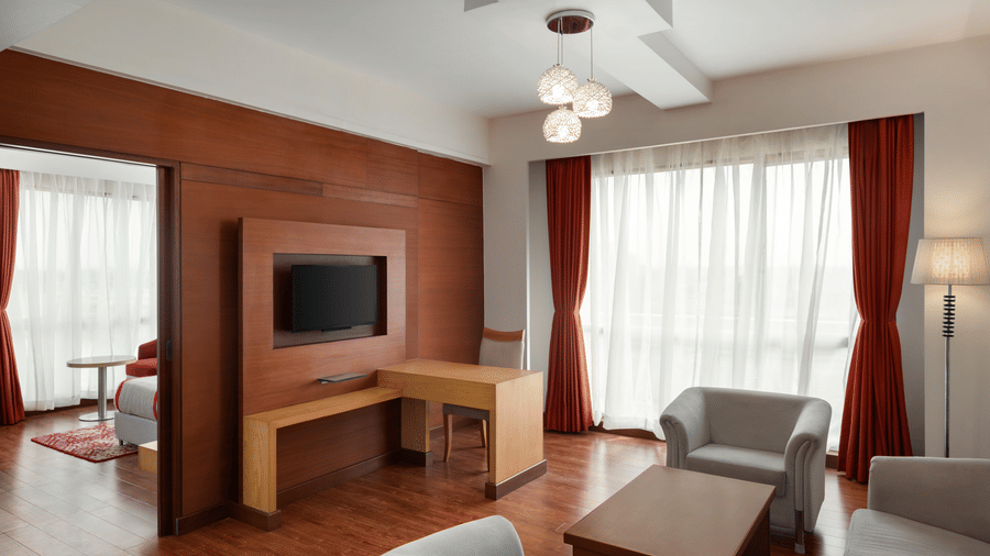 Living area of the King Suite at Ramada by Wyndham Bodh Gaya Mahabodhi featuring a television, multiple sofas, a wooden table in the centre, and elegant wooden furnishings.