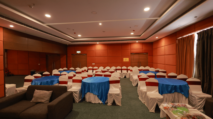 A function room is set up with round tables, blue and red linen, and a small sofa lounge area at Ramada by Wyndham Bodh Gaya Mahabodhi.
