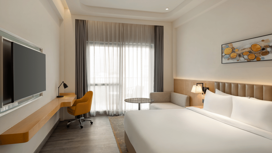 Wide-angle view of a contemporary hotel room with a king-size bed, flat-screen TV, work desk with chair, and large curtained windows.