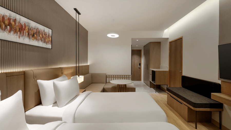 Spacious twin-bed hotel room with white linens, wooden furniture, recessed ceiling lighting, and a wall-mounted flat-screen television.
