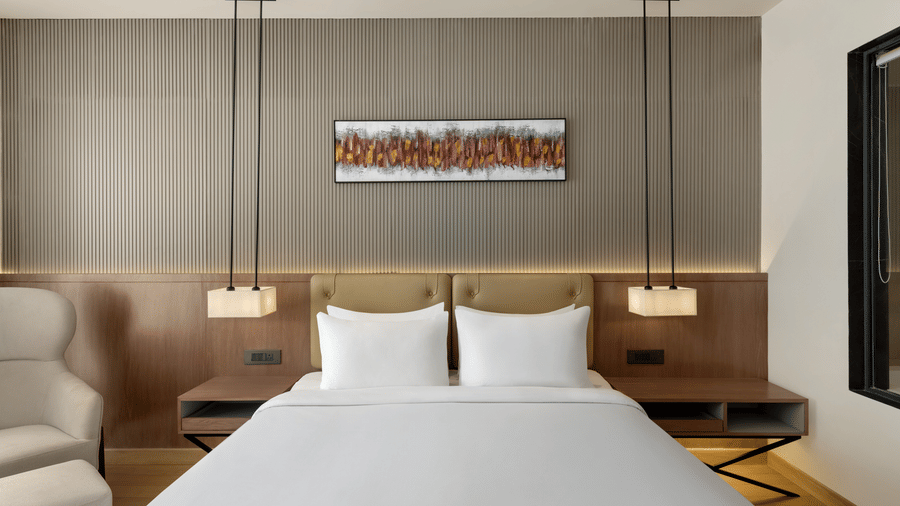 Elegant hotel room featuring a king-size bed with white linens, pendant bedside lighting, warm wooden tones, and a vibrant abstract wall painting.