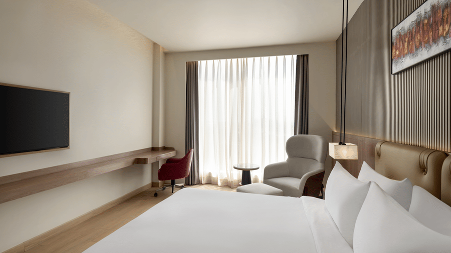 ontemporary hotel room at Ramada by Wyndham Sonipat Murthal, with a wide king-size bed, wooden platform base, flat-screen TV on a low console, and a cushioned accent chair by the window.