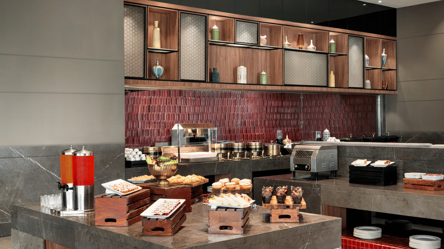 Close-up of a buffet counter station with beverage dispensers, food trays, and rich wooden cabinetry in a deep burgundy-toned setting.