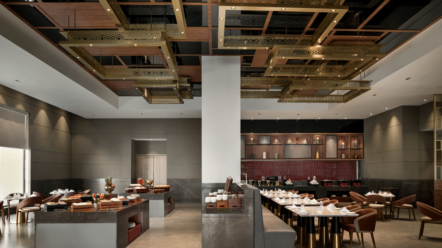 Modern restaurant interior with an exposed industrial ceiling, gold geometric framework, and neatly arranged dining tables under pendant lighting.